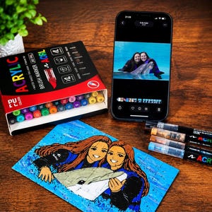 Pode incluir: Um conjunto de marcadores acrílicos de ponta de pincel numa caixa preta com tampas coloridas, um smartphone a exibir uma fotografia de duas pessoas com um golfinho e uma ilustração feita à mão da mesma cena num fundo azul com três marcadores.