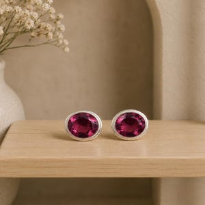 May include: A pair of oval-shaped stud earrings featuring deep magenta-colored faceted stones set in a smooth, polished silver-toned metal frame. The earrings are displayed side-by-side, showcasing the vibrant pinkish-red hue and the reflective quality of the cut gemstone surfaces.