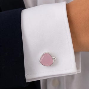 Puede incluir: Un gemelo de plata con una piedra lisa de color rosa pálido, de forma triangular y bordes redondeados. El gemelo está sujeto a través de los ojales del puño de una camisa blanca de manga larga, que se lleva debajo de una americana azul marino.