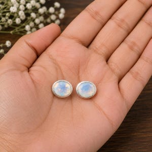 May include: A pair of oval-shaped stud earrings featuring iridescent, milky white cabochon stones set in polished silver-toned metal bezels. The stones display subtle blue and pink flashes when catching the light, creating a soft, glowing appearance.