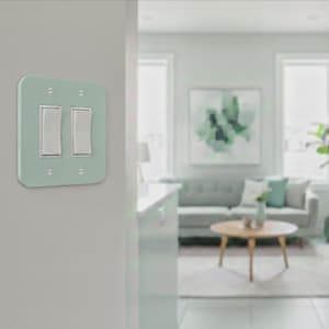 May include: A light sage green double light switch wall plate featuring two white rocker switches. The rectangular plate is secured to the wall with four visible silver screws, one in each corner, providing a clean and functional electrical fixture.