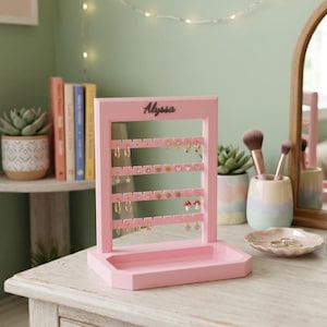 May include: A light pink wooden earring holder with a rectangular base and four horizontal bars featuring pre-drilled holes for jewelry storage. The top frame displays the name Alyssa in black script lettering, and the stand is empty.