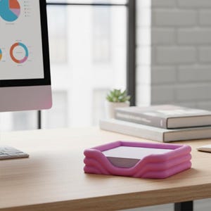 May include: A pink desk organizer with a wavy design holds a stack of white notepaper. The organizer sits on a light-colored wooden desk, with a computer monitor and books in the background. The office setting is bright and modern.