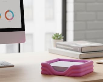 May include: A pink desk organizer with a wavy design holds a stack of white notepaper. The organizer sits on a light-colored wooden desk, with a computer monitor and books in the background. The office setting is bright and modern.