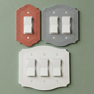 May include: Three decorative wall switch plates with a scalloped edge design. One single-gang plate in terracotta, one double-gang plate in gray, and one triple-gang plate in white. Each plate features white rocker switches and visible metal screws.