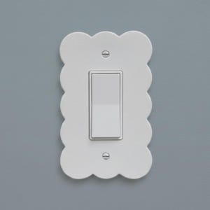 May include: A white light switch with a scalloped edge plate. The switch is rectangular and centered on the plate. Two screws secure the plate to the wall. The background is a solid light gray.