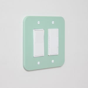 May include: A mint green light switch plate with two white switches. The plate is square with rounded corners and has four small screws. The switches are a standard toggle and a dimmer.