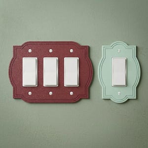 May include: A dark red triple light switch plate with a textured finish and three white rocker switches, alongside a smaller light green single light switch plate with a smooth finish and one white rocker switch, both featuring decorative edges.