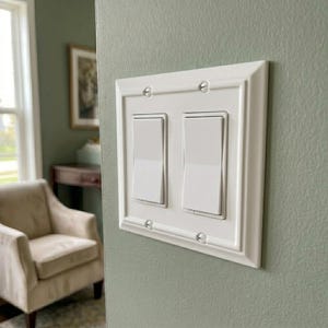 May include: A white, rectangular light switch plate with two rocker switches. The plate has a decorative, framed edge and is mounted on a light green wall. The switches are in the on position.