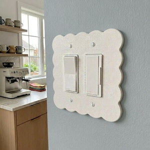 May include: A white, scalloped-edge light switch cover with two light switches. The switches are rectangular and silver. The cover is mounted on a light blue wall. The kitchen is visible in the background.