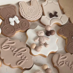 Peut inclure: Une collection de biscuits décorés aux formes variées, incluant des oursons, des bavoirs, des bodies et des plaques festonnées avec le mot Baby en écriture cursive. Les biscuits sont décorés avec des glaçages dans les tons beige, brun et blanc.
