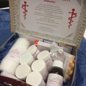 May include: A gift box containing a collection of small white containers, a large white pillar candle, a brown glass bottle, and a small clear box of yellow and pink earplugs. The inside lid features a printed card with Norwegian text and red medical symbols.