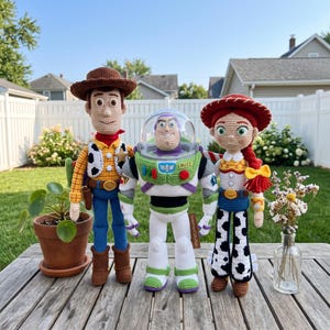 May include: Three handmade crochet dolls representing characters with a cowboy in a brown hat, a space ranger with a clear helmet, and a cowgirl with red yarn hair. Each doll features detailed stitching, vibrant colors, and distinct outfits.