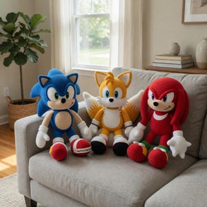 May include: Three handmade crochet plush toys featuring Sonic the Hedgehog in blue, Tails in yellow with two tails, and Knuckles in red. Each character has white accents, large eyes, and stitched details. One foot on the blue plush has a small white tag.
