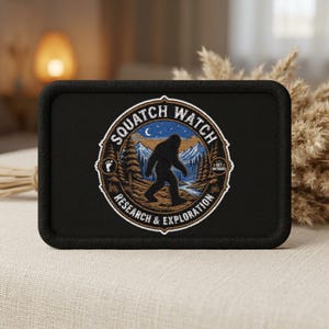May include: A rectangular black patch featuring an embroidered circular emblem. The design shows a silhouette of a creature walking through a forest with mountains and a crescent moon in the background. Text reads SQUATCH WATCH RESEARCH & EXPLORATION.