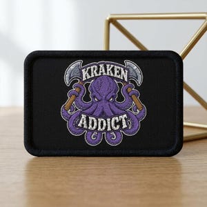 Op de afbeelding: Een rechthoekige zwarte geborduurde patch met een paarse octopus met een strenge uitdrukking, die twee zilveren bijlen met bruine handvatten vasthoudt. De tekst KRAKEN staat in een boog boven de octopus en het woord ADDICT staat eronder in wit.