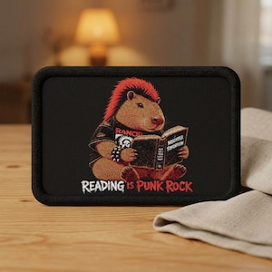 May include: A rectangular black patch featuring an embroidered capybara with a red mohawk, wearing a black Rancid band t-shirt and a spiked wristband. The capybara is reading a book titled Brighter Tomorrow. The text Reading Is Punk Rock is below.