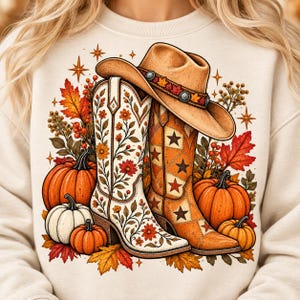 Könnte beinhalten: Ein cremefarbenes Sweatshirt mit einem Grafikdruck eines beigen Cowboyhuts, der auf zwei Cowboystiefeln ruht. Ein Stiefel ist weiß mit Blumenmustern, der andere orange mit Sternmotiven. Umgeben von orangefarbenen Kürbissen und Herbstblättern.