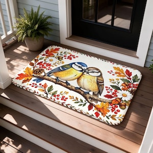Puede incluir: Un felpudo rectangular con fondo color crema y una ilustración central de dos pájaros pequeños posados en una rama. El diseño incluye hojas otoñales en tonos naranja, rojo y amarillo, junto con margaritas blancas y bayas rojas.