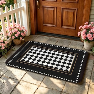 May include: A rectangular doormat featuring a central black and white chequered pattern bordered by a thin gold line and a black frame with a white dotted edge. The mat is positioned on a stone patio in front of a wooden door with pink flowers.