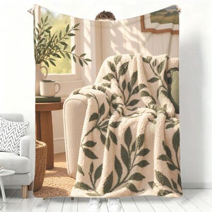May include: A cream-colored sherpa fleece throw blanket featuring a repeating dark green leaf pattern. The blanket is draped over a sofa in a room with a wooden side table, a white mug, and a potted plant visible in the background scene printed on it.