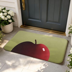 Puede incluir: Un felpudo rectangular con fondo verde claro y un gráfico grande y estilizado de una manzana de color rojo oscuro centrado en la superficie. El diseño de la manzana incluye un pequeño tallo marrón y un reflejo blanco en la fruta.