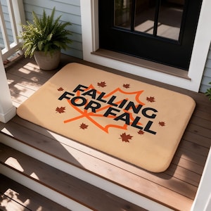 Puede incluir: Un felpudo rectangular de color tostado con el texto negro FALLING FOR FALL centrado sobre el contorno de una hoja de arce naranja grande, rodeado de varios iconos de hojas de arce marrón oscuro dispersos por la superficie.