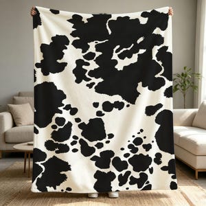 May include: A rectangular throw blanket featuring a high-contrast black and off-white cow print pattern. The fabric displays irregular, organic black spots scattered across a solid off-white background, covering the entire surface of the textile.