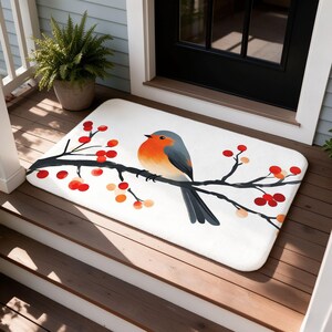 May include: A rectangular white doormat featuring a graphic illustration of a robin perched on a black tree branch. The branch is adorned with clusters of bright red and orange berries, extending across the center of the mat surface.