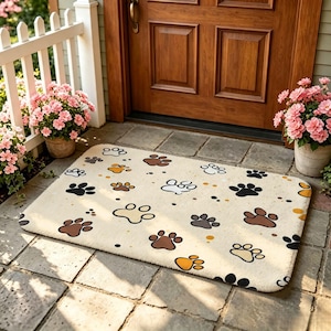 May include: A rectangular doormat with a cream-colored background featuring a repeating pattern of animal paw prints in various shades of brown, black, and tan, interspersed with small solid dots in matching colors across the entire surface.