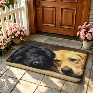 Puede incluir: Un felpudo rectangular que presenta una ilustración realista y detallada de dos perros Labrador retriever descansando uno al lado del otro. Un perro tiene un pelaje negro sólido, mientras que el otro tiene un pelaje amarillo. Ambos se ven.
