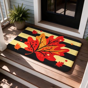 Puede incluir: Un felpudo rectangular con un patrón de rayas horizontales negras y amarillas. El centro de la alfombra muestra una hoja de otoño grande y detallada en tonos rojos, naranjas y amarillos, acompañada de motivos de hojas más pequeños.
