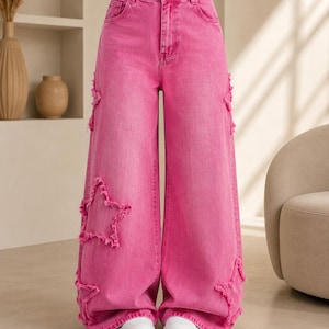 May include: High-waisted wide-leg jeans in a vibrant hot pink wash, featuring a button and zip fly closure and multiple frayed-edge star-shaped patches stitched onto the legs. The denim trousers have a relaxed fit and a classic five-pocket design.