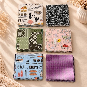 May include: Six stacks of square fabric swatches with pinked edges. Designs include a cream background with black cat and bakery illustrations, a black floral print, a green patchwork pattern, a pink floral print with a red car, a light blue bakery print, and a purple gingham check.