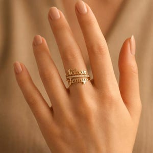 Op de afbeelding: Een goudkleurige ring met twee gestapelde naamplaatjes met de namen Nelson en Jenny in een cursief lettertype, gescheiden door een dunne band ingelegd met kleine heldere steentjes. Het metaal heeft een gepolijste afwerking en een warme tint.