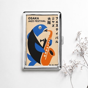 May include: A rectangular silver metal cigarette case featuring a graphic print of a blue and black fish wearing a tuxedo and playing an orange saxophone. The text reads OSAKA JAZZ FESTIVAL and 10.10.1991 with Japanese characters included.