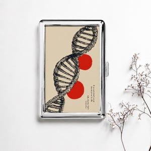 May include: A rectangular silver metal case featuring a beige insert with a black ink illustration of a DNA double helix. Two solid red circles are positioned behind the helix, and vertical Japanese text is printed in the lower right corner.