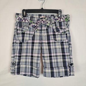 May include: Gray plaid shorts with a floral patterned belt and bow. The shorts have front pockets, button details on the cuffs, and are hanging on a black hanger. The plaid pattern includes black, white, and purple lines.