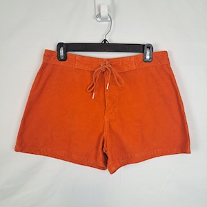 May include: Bright orange corduroy shorts with a drawstring waist. The shorts are hanging on a black hanger against a white background. The shorts have a classic, casual style.