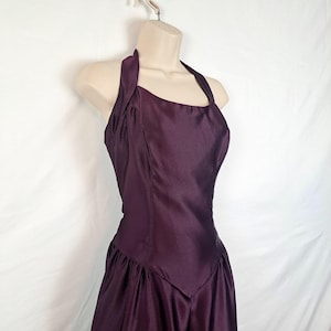 May include: A plum-colored halter dress with a satin finish. The dress features a fitted bodice and a flared skirt. The halter straps are wide and the neckline is square. The dress is displayed on a mannequin against a white background.