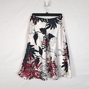 May include: A knee-length satin skirt featuring a white base with a bold floral print. The pattern consists of large, stylized chrysanthemum flowers in deep black and dark red, accented by fine black line-drawn leaves and stems.
