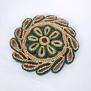 May include: A round, woven coaster with a floral design. The center features a flower with green petals and a dark brown center. The outer ring has a leaf-like pattern in green and natural tones. The coaster is made of natural fibers.
