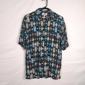 May include: A short-sleeved button-down shirt with a geometric pattern in shades of blue, gray, and beige. The shirt has a collar and a button-up front.
