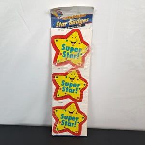 May include: A package of "Star Badges" awards to wear. The badges are yellow stars with red borders and the words "Super Star!" in blue. The package contains 24 awards.