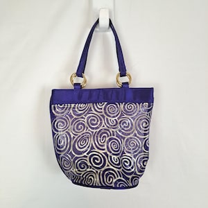 May include: A purple tote bag with gold-colored handles and a silver swirl pattern. The bag has a rectangular shape with a rounded bottom and is suspended from a white hook. The handles are attached to the bag with gold-colored rings.