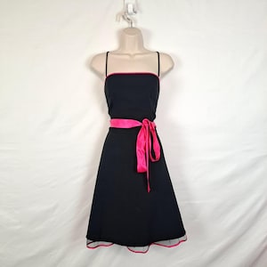 May include: Black dress with a pink ribbon belt and trim. The dress has a square neckline with thin straps and a pink trim. The skirt is A-line, and the dress is displayed on a mannequin.