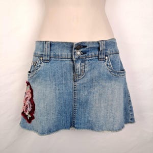 May include: A light blue denim mini skirt with a frayed hem. The skirt has a floral embroidery design on the left side pocket.