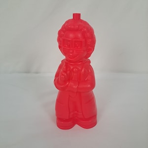 May include: A solid red plastic figurine of a bespectacled person in a lab coat, with a small spout on top. The figure has a smiling face and crossed arms, and is standing on a white surface.