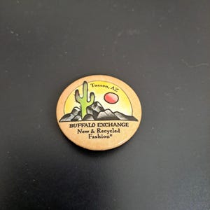 May include: A round button with a desert scene featuring a cactus, mountains, and a red sun. The text "Buffalo Exchange New & Recycled Fashion" is printed on the button, along with the location "Tucson, AZ".