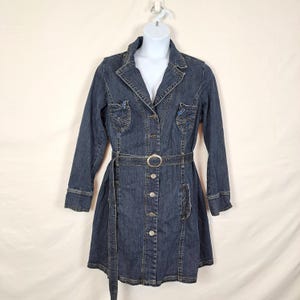 May include: A dark blue denim jacket dress featuring a collared neckline, long sleeves with cuffs, and a front button-down closure. The garment includes two chest pockets with decorative stitching, a matching fabric belt with a silver buckle.